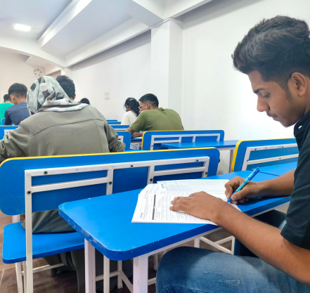 A group of students seated in a well-lit classroom at blue desks, focused on writing their exams.